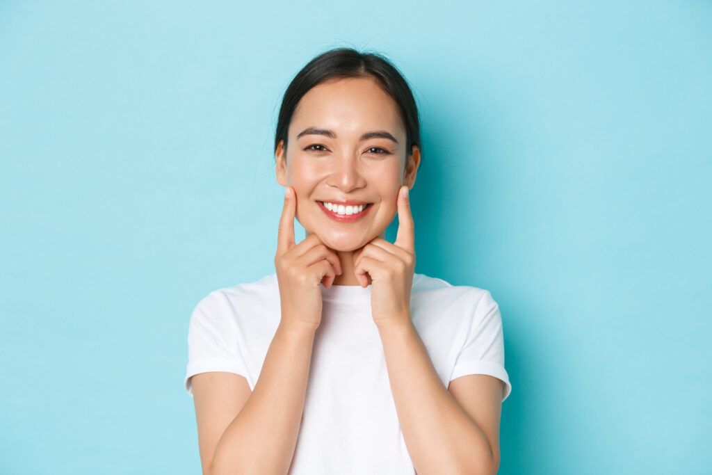 close up of beautiful asian girl looking pleased, smiling white teeth, touching cheeks, recommend skincare product, cosmetics for facial skin, standing light blue background